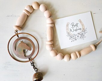 May include: A wooden bead garland with a metal horse head charm and a small bell. A card with a horseshoe and the words "Best Wishes" is also present. The items are arranged on a white surface.