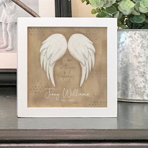 May include: White framed picture with a beige background featuring a pair of white angel wings. The text "I am with you always" is written in cursive script below the wings. The name "Jenny Williams" and the dates "1922 - 2020" are written below the text.