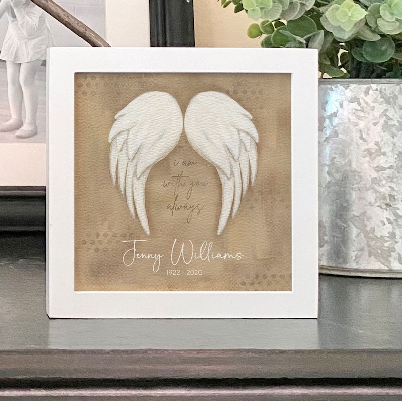 May include: White framed picture with a beige background featuring a pair of white angel wings. The text "I am with you always" is written in cursive script below the wings. The name "Jenny Williams" and the dates "1922 - 2020" are written below the text.