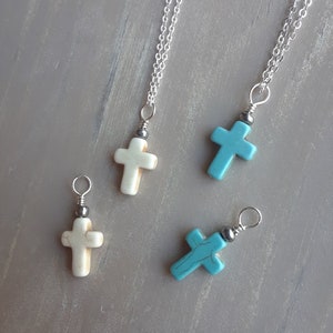 May include: Four cross pendant necklaces. Two are off-white, and two are turquoise. Each cross is attached to a silver chain. The pendants have a small silver bead above the cross.