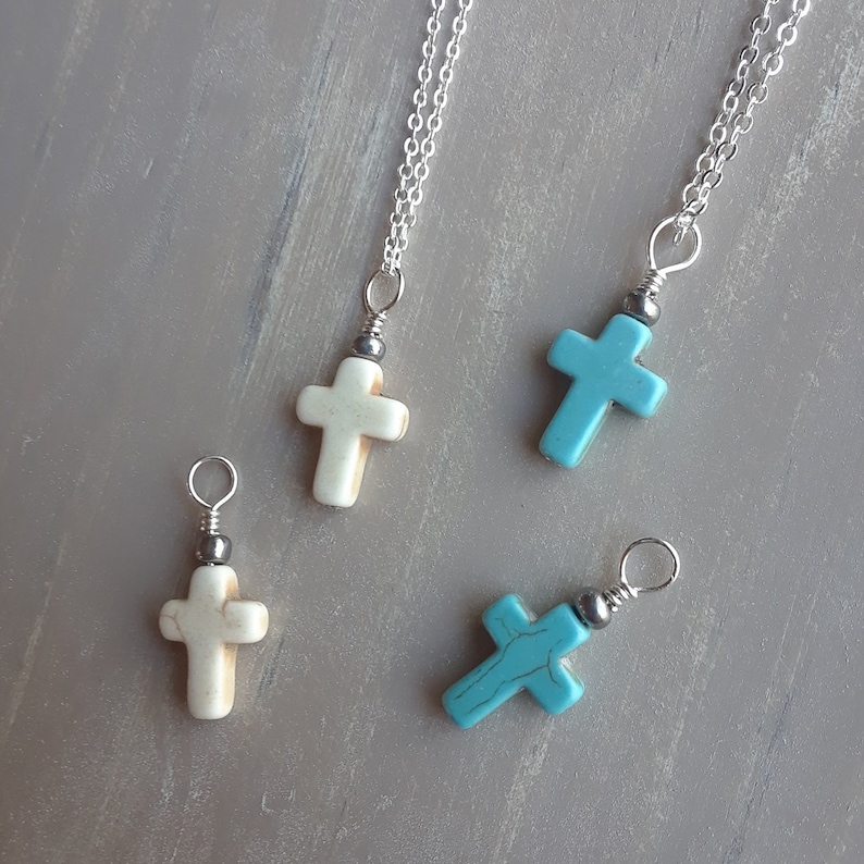 May include: Four cross pendant necklaces. Two are off-white, and two are turquoise. Each cross is attached to a silver chain. The pendants have a small silver bead above the cross.