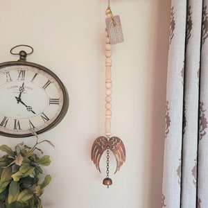 May include: A wooden bead garland with a metal angel wing wind chime and a bell at the bottom. The garland is hanging from a hook on a white wall. The text on the tag reads "Bell Wind Chime".