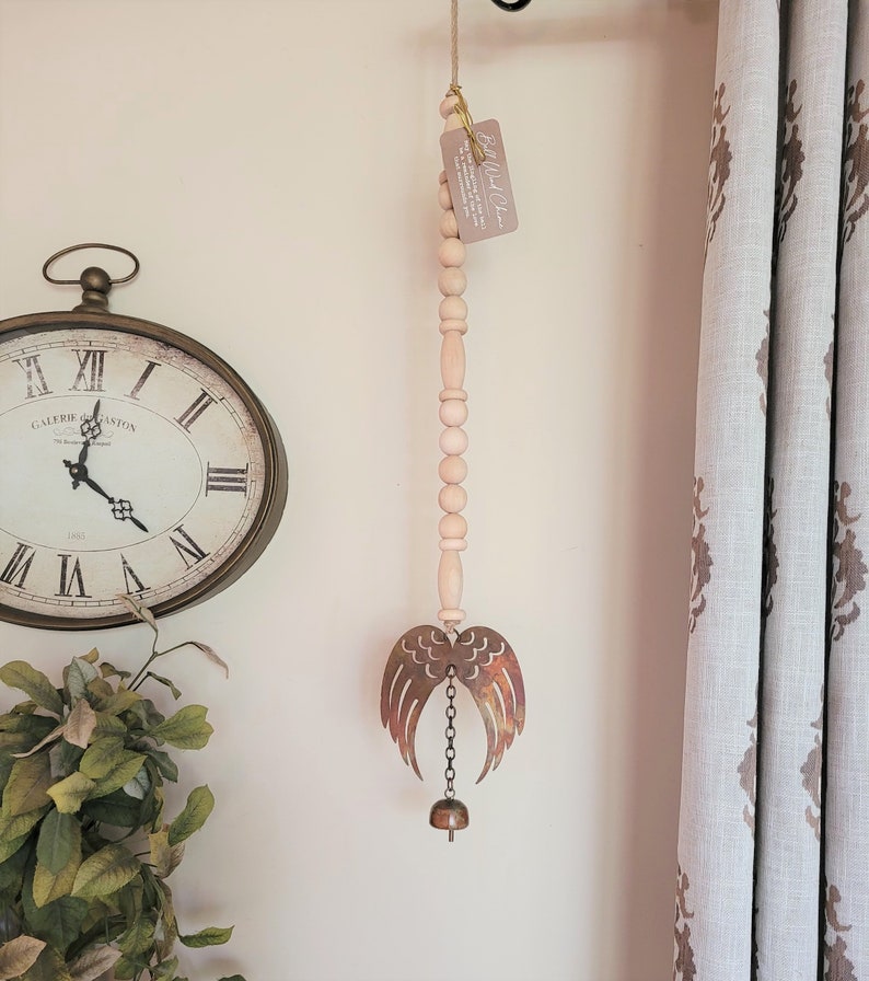 May include: A wooden bead garland with a metal angel wing wind chime and a bell at the bottom. The garland is hanging from a hook on a white wall. The text on the tag reads "Bell Wind Chime".