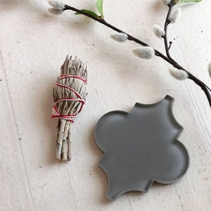 May include: A bundle of dried sage tied with red string, and a gray ceramic tile with a decorative design, on a white wooden surface.