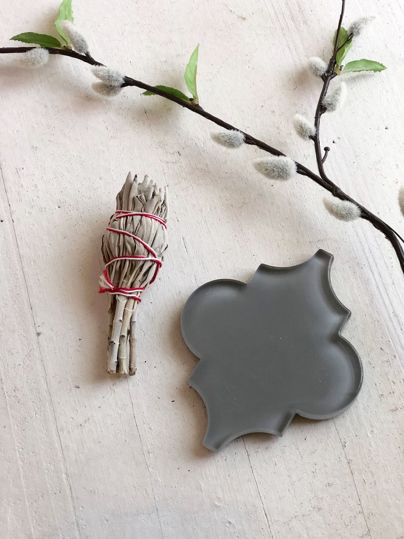 May include: A bundle of dried sage tied with red string, and a gray ceramic tile with a decorative design, on a white wooden surface.