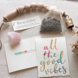 May include: A gift box filled with items for good vibes, including a bell wind chime, a small bag of lavender and rose petals, a pink crystal, and a card with the text "all the good vibes".