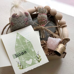 May include: A small gift box filled with items for a housewarming gift. The box contains a sage smudge stick, a small pink crystal, a bell, a bag of dried lavender and rose petals, and a card with the word "home" on it.