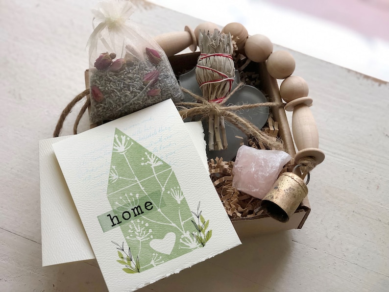 May include: A small gift box filled with items for a housewarming gift. The box contains a sage smudge stick, a small pink crystal, a bell, a bag of dried lavender and rose petals, and a card with the word "home" on it.