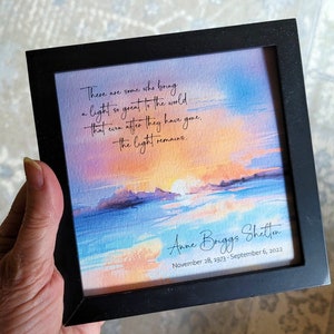 May include: A framed watercolor print with a black frame. The artwork depicts a sunset over water, with text that reads "There are some who bring a light so great to the world...the light remains." The print is signed "Anne Briggs Shelton" with dates.