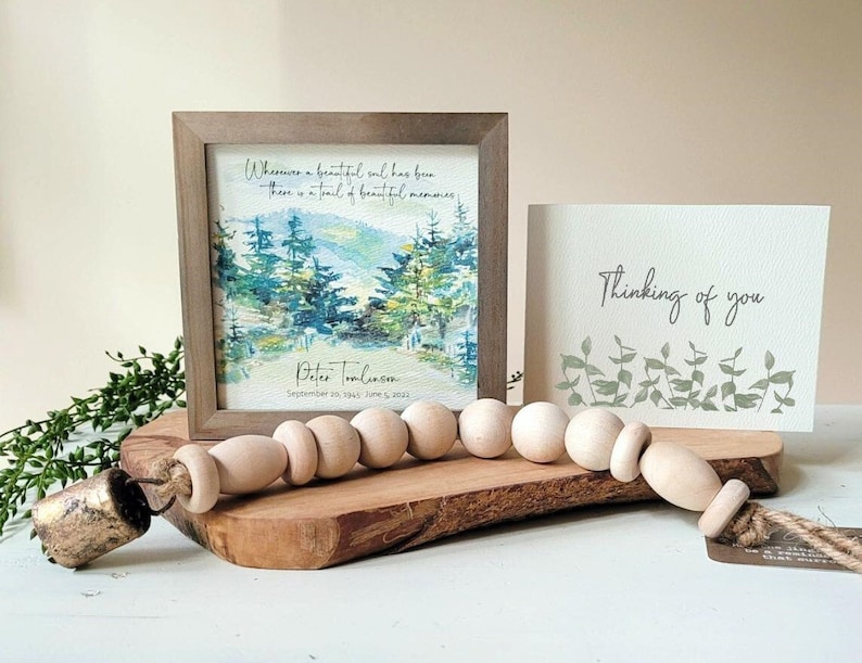 May include: A framed watercolour painting of a forest scene with the quote "Wherever a beautiful soul has been, there is a trail of beautiful memories."  The frame is brown and the painting is on a white background. The text "Peter Tomlinsom, September 20, 1945 - June 5, 2022" is printed below the painting. A small card with the text "Thinking of you" is also visible in the image.