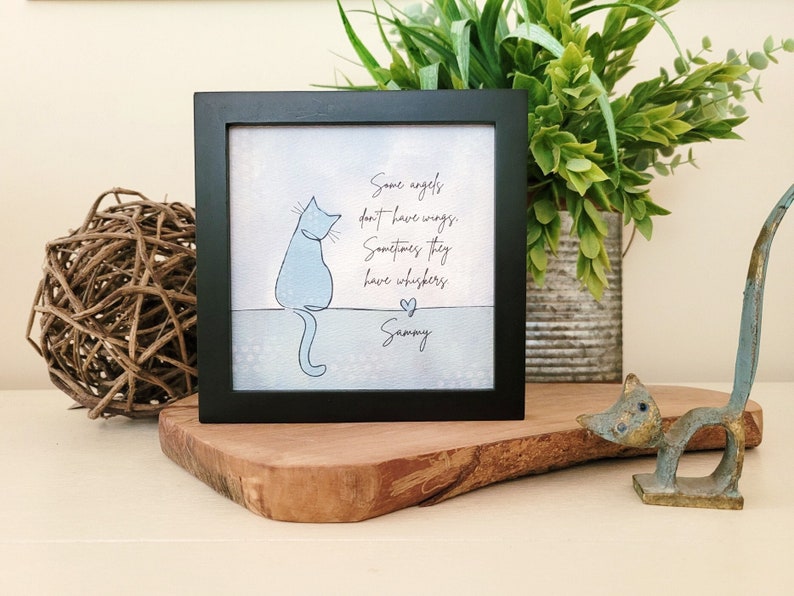May include: A framed print with a black border features a light blue cat illustration and the text "Some angels don't have wings. Sometimes they have whiskers. Sammy". The print is displayed on a wooden surface.