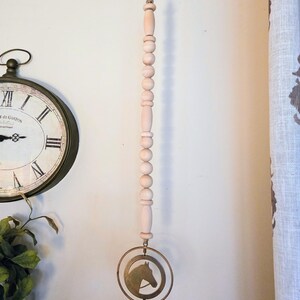 May include: A decorative wind chime featuring a series of light pink wooden beads suspended from a jute rope. A brass-colored ring with a horse head silhouette hangs below, with a small bell at the bottom. The chime is designed for indoor use.