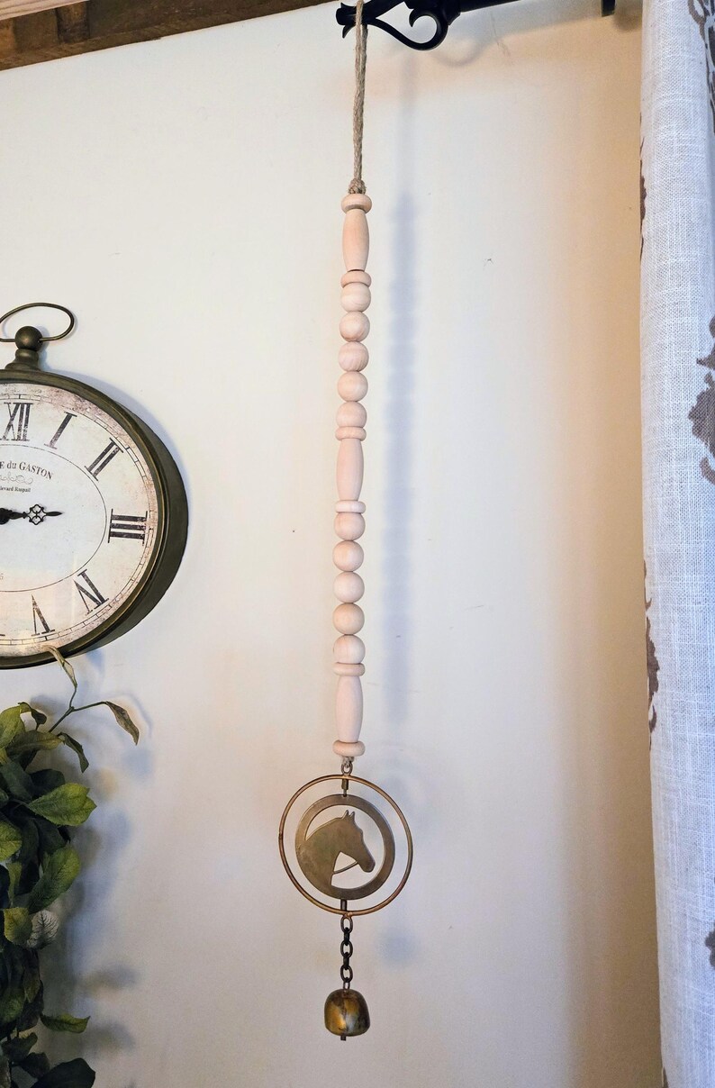May include: A decorative wind chime featuring a series of light pink wooden beads suspended from a jute rope. A brass-colored ring with a horse head silhouette hangs below, with a small bell at the bottom. The chime is designed for indoor use.