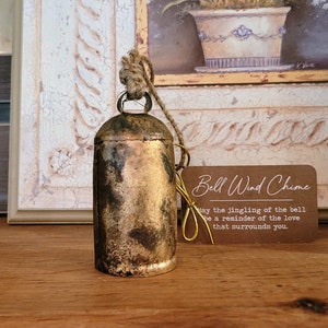 May include: A vintage-style bell wind chime with a tarnished gold finish, hanging from a natural rope. A small tag with the text "Bell Wind Chime" and a message is attached. The bell is set against a backdrop of a framed painting.