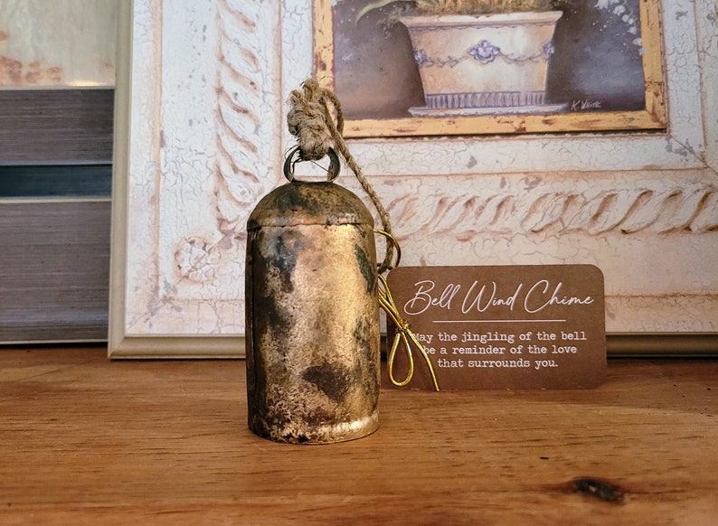 May include: A vintage-style bell wind chime with a tarnished gold finish, hanging from a natural rope. A small tag with the text "Bell Wind Chime" and a message is attached. The bell is set against a backdrop of a framed painting.