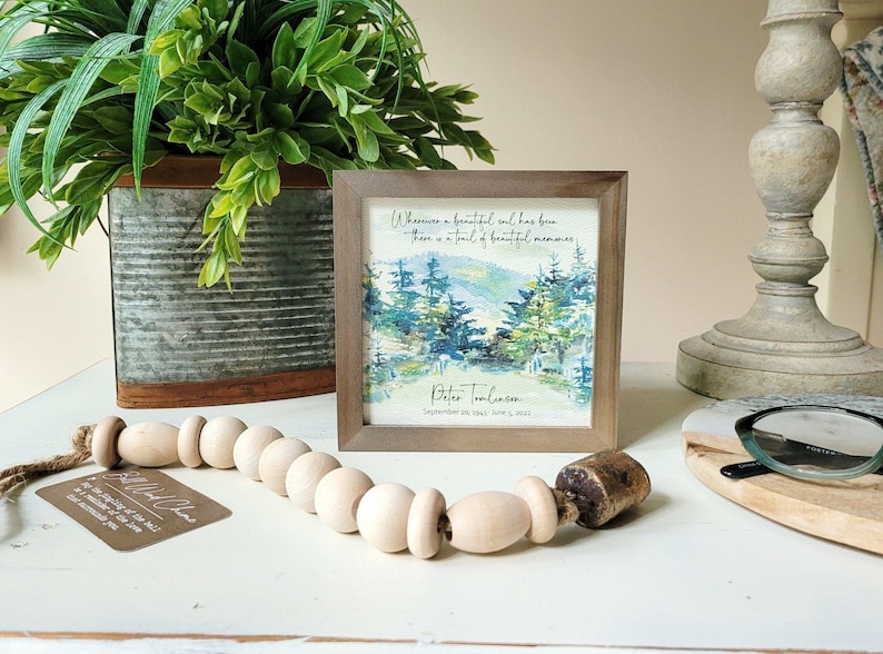 May include: A framed watercolour painting of a forest scene with a quote about beautiful souls leaving behind beautiful memories. The quote reads "Wherever a beautiful soul has been, there is a trail of beautiful memories." The frame is brown wood and the painting is signed "Peter Tomlinson" with the date "September 18th 1941 - June 5, 2023".