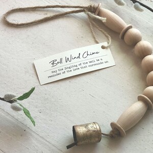 May include: A wooden bead wind chime with a small gold bell. The chime is made of natural wood beads and has a simple, rustic design. The tag attached to the chime reads "Bell Wind Chime May the jingling of the bell be a reminder of the love that surrounds us."