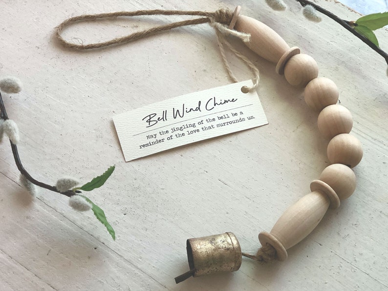 May include: A wooden bead wind chime with a small gold bell. The chime is made of natural wood beads and has a simple, rustic design. The tag attached to the chime reads "Bell Wind Chime May the jingling of the bell be a reminder of the love that surrounds us."