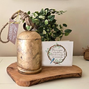 May include: A gold-colored metal bell with a rope handle, sitting on a wooden cutting board. A card with the text "May the ringing of the bell be a reminder of the joy that surrounds you" is next to the bell.