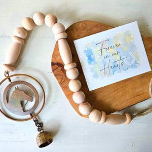 May include: A wooden bead garland with a metal horse head ornament and a small bell. A card with the text "Forever in our Hearts" rests on a wooden board. The garland is made of natural wood beads.