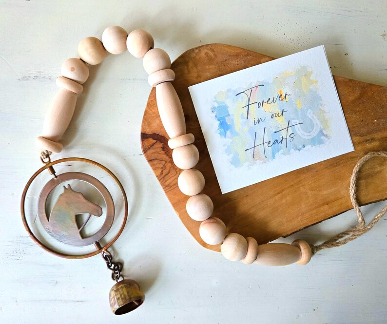 May include: A wooden bead garland with a metal horse head ornament and a small bell. A card with the text "Forever in our Hearts" rests on a wooden board. The garland is made of natural wood beads.