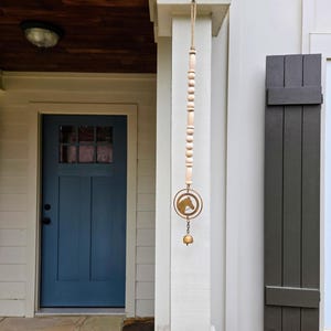 May include: A decorative wind chime with a horse head silhouette, hanging from a wooden and beaded strand. The chime is suspended from a porch column next to a blue door and dark gray shutters.