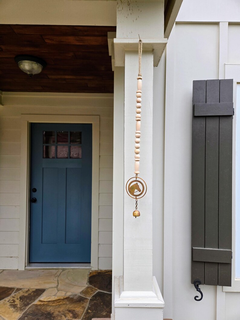 May include: A decorative wind chime with a horse head silhouette, hanging from a wooden and beaded strand. The chime is suspended from a porch column next to a blue door and dark gray shutters.