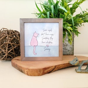 May include: A framed print featuring a pink cat illustration and the text "Some angels don't have wings, Sometimes they have whiskers, Sammy." The frame is brown, and the artwork is displayed on a wooden surface.