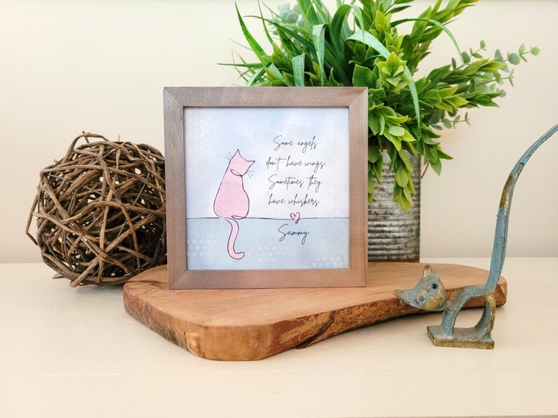 May include: A framed print featuring a pink cat illustration and the text "Some angels don't have wings, Sometimes they have whiskers, Sammy." The frame is brown, and the artwork is displayed on a wooden surface.
