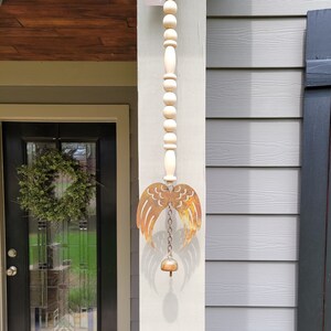 May include: A rustic metal angel wing wind chime with a wooden bead chain and a bell at the bottom. The wind chime is hanging from a porch.