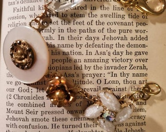 May include: A close-up of a book with a white button and gold accents. The book is open to a page with text about the Bible. The button is surrounded by other buttons and beads, some of which are white, gold, and clear.