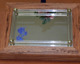 May include: A wooden picture frame with a glass insert and gold-colored accents. The frame has a mirror inside with a blue silhouette of a unicorn and a pink flower.