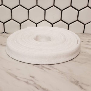 May include: A white cotton twill tape rolled up into a circle. The tape is approximately 1 inch wide.