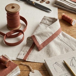 May include: A still-life image of sewing and tailoring supplies. Includes a shirt collar with rust-colored trim, scissors, spools of thread, wooden buttons, fabric swatches, and a notebook with sketches. The color palette is neutral with rust accents.