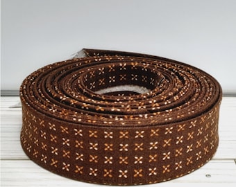May include: A rolled-up brown ribbon with a repeating pattern of small white and tan crosses. The ribbon is made of fabric and appears to be new. The background is a light gray and white.