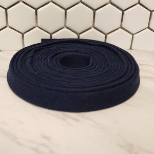 May include: A rolled length of navy blue bias tape. The tape is coiled tightly, forming a circular shape. The fabric has a soft, matte appearance. The background shows a white hexagonal tile pattern and a white marble surface.