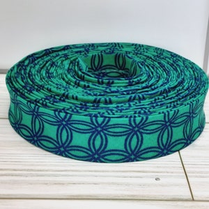 May include: A tightly coiled roll of green fabric ribbon with a repeating navy blue interlocking circle pattern. The ribbon is displayed on a light-colored wooden surface, showcasing its design and texture.