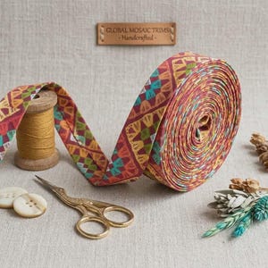 May include: A roll of patterned ribbon with a geometric design in shades of red, yellow, and teal. A wooden spool of thread, gold scissors, two buttons, and dried flowers are also present. The sign reads "GLOBAL MOSAIC TRIMS - Handcrafted".