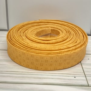 May include: A roll of yellow bias tape with a repeating floral pattern. The tape is tightly coiled, revealing the edges and the detailed design. The tape is made of fabric and is suitable for sewing and crafting projects.