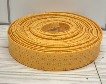 May include: A roll of yellow bias tape with a repeating floral pattern. The tape is tightly coiled, revealing the edges and the detailed design. The tape is made of fabric and is suitable for sewing and crafting projects.