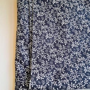 May include: A folded piece of dark navy blue fabric featuring a repeating white botanical pattern of delicate vines, small leaves, and tiny flowers. The textile has a visible woven texture and a lighter, off-white underside visible at the edge.