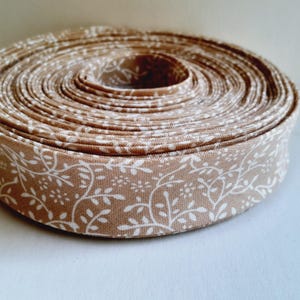 May include: A coiled roll of tan fabric bias tape featuring a repeating white botanical pattern of delicate vines, leaves, and small flowers. The textile ribbon is wound into a tight, circular spool, showcasing the continuous floral print.