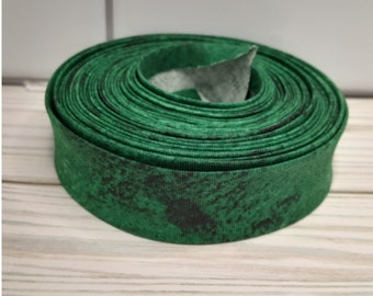 May include: A roll of green fabric bias tape with a dark, mottled pattern. The tape is tightly wound, revealing a glimpse of the lighter-colored backing. The fabric appears to be cotton or a similar material, suitable for sewing and crafting projects.