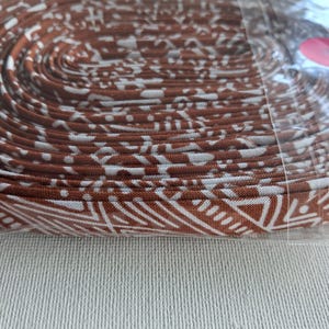 May include: A coiled length of fabric trim featuring a repeating geometric pattern in white against a rich brown background. The trim is neatly wound in a circular shape and encased in a clear plastic sleeve, showing a consistent woven texture.