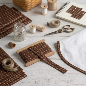 May include: A collection of sewing supplies including brown fabric with a white cross pattern, a wooden spool of matching ribbon, a glass jar of pins, a thimble, white thread spools, a measuring tape, scissors, and a white cloth with brown trim.