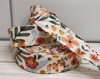 May include: A roll of floral fabric ribbon featuring a cream background with a repeating pattern of orange, red, and yellow flowers, and green leaves. The ribbon is rolled up and partially unrolled, showing the detailed floral design.