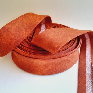 May include: A roll of burnt orange bias tape featuring a textured, mottled fabric surface. The tape is wound into a circular coil with one end extending outward, revealing a white, adhesive-like backing along the center of the strip.