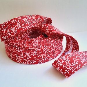 May include: A continuous roll of red fabric ribbon featuring a repeating white botanical pattern of small leaves and flowers. The ribbon is coiled into a circular shape with one end extending outward, showcasing the consistent print on the textile.