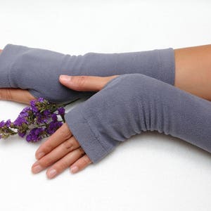 May include: A pair of gray fingerless gloves with a ribbed texture. The gloves are worn on a person's hands, and a small bouquet of purple flowers is held in the right hand.