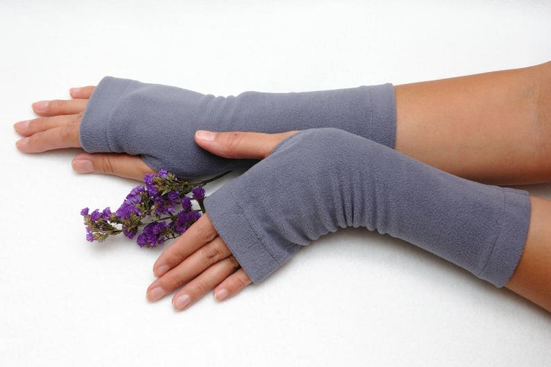 May include: A pair of gray fingerless gloves with a ribbed texture. The gloves are worn on a person's hands, and a small bouquet of purple flowers is held in the right hand.